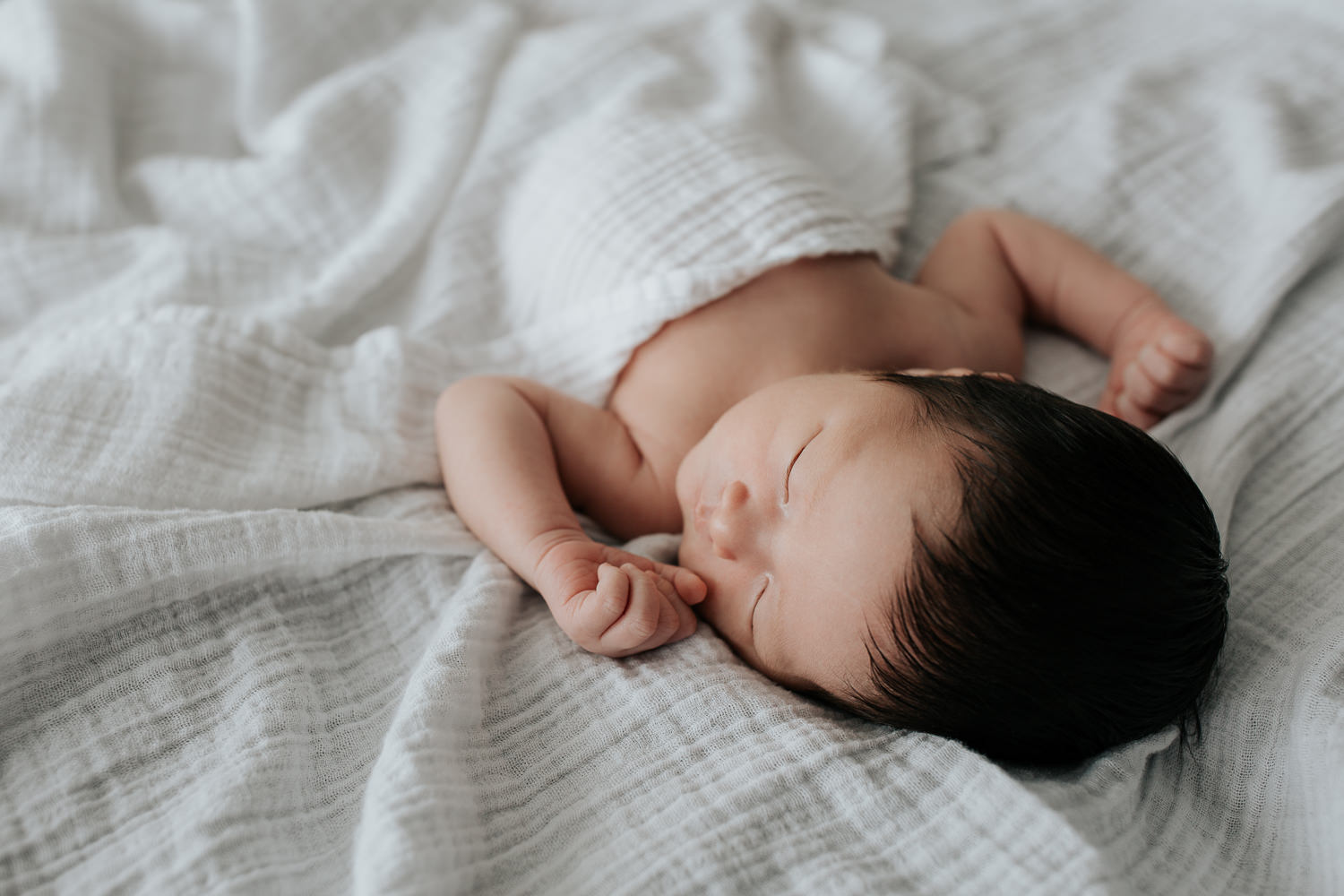 2 week old baby boy with long dark hair in diaper lying on bed, body covered by white swaddle, arms out, head to side, sleeping - Stouffville Lifestyle Photography