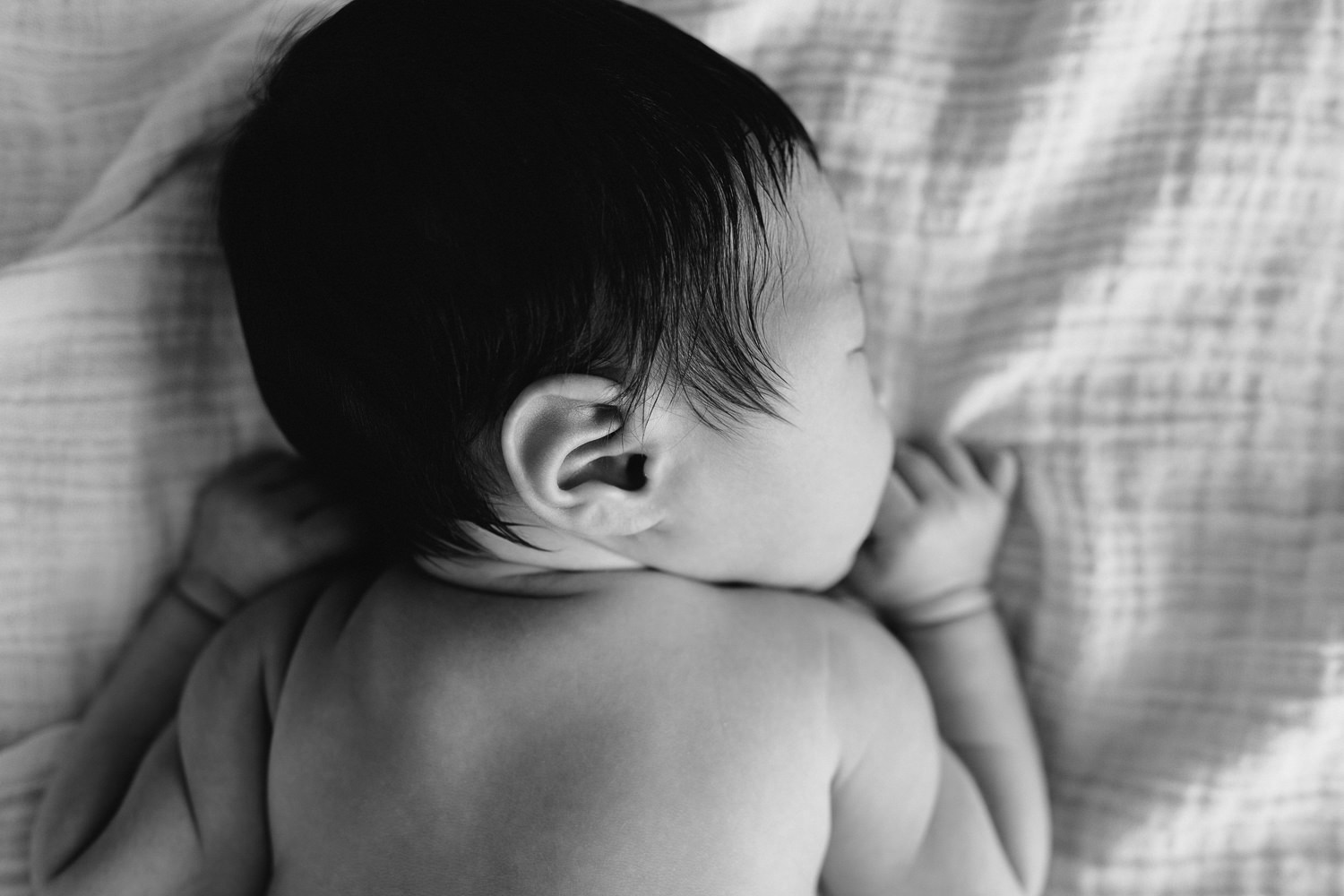 2 week old baby boy with long dark hair in diaper lying on stomach, arms up by face, legs tucked in, head to side, sleeping on bed, close up of ear - Stouffville In-Home Photography