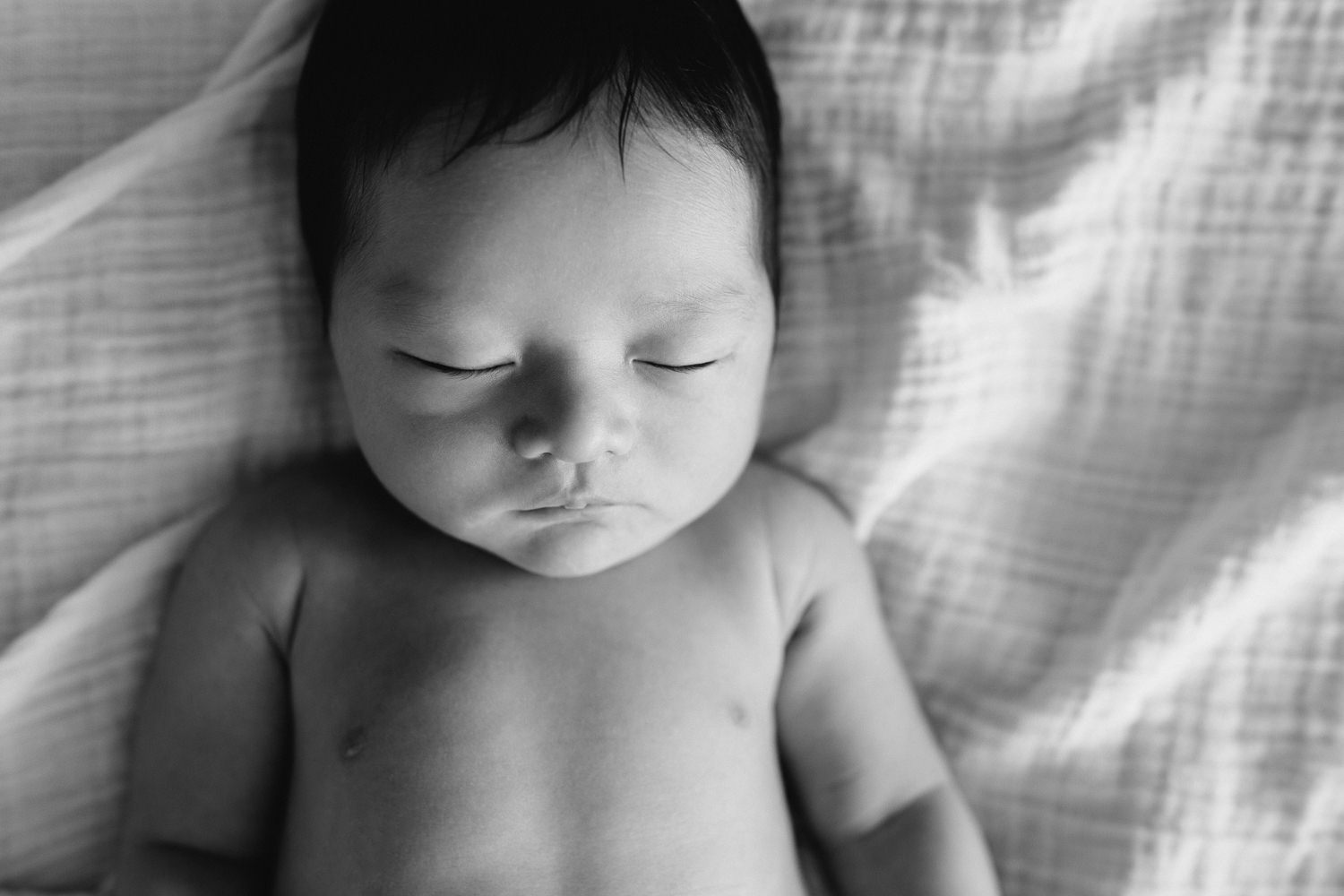 2 week old baby boy with long dark hair in diaper lying on back, hand resting on stomach, blanket over body, sleeping on bed, close up of face - Uxbridge In-Home Photography