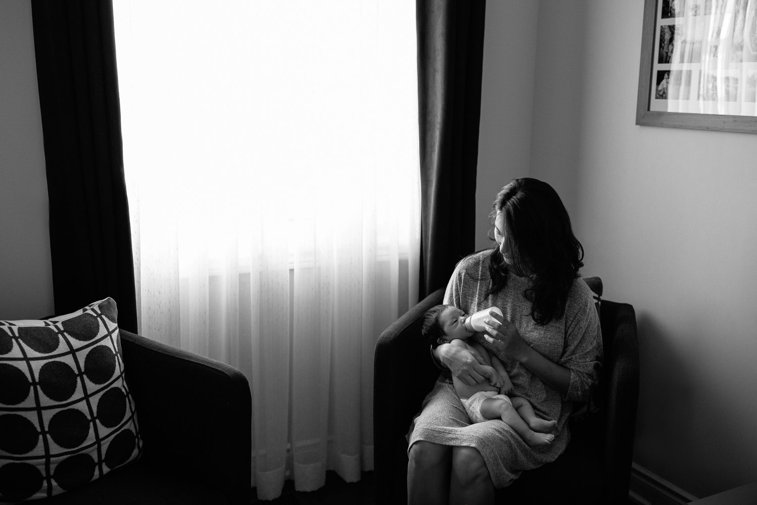 mom with long dark hair sits in armchair next to window feeding 2 week old baby boy in diaper a bottle - Stouffville In-Home Photos