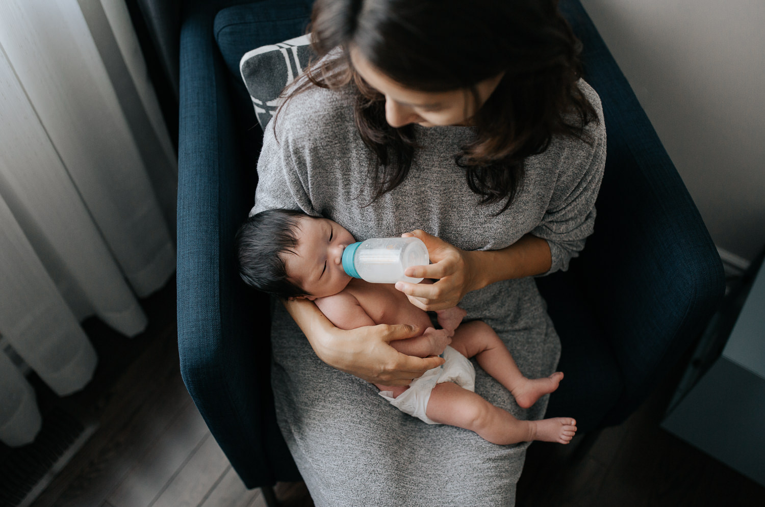 mom with long dark hair sits in blue armchair feeding 2 week old baby boy in diaper a bottle - Uxbridge In-Home Photos