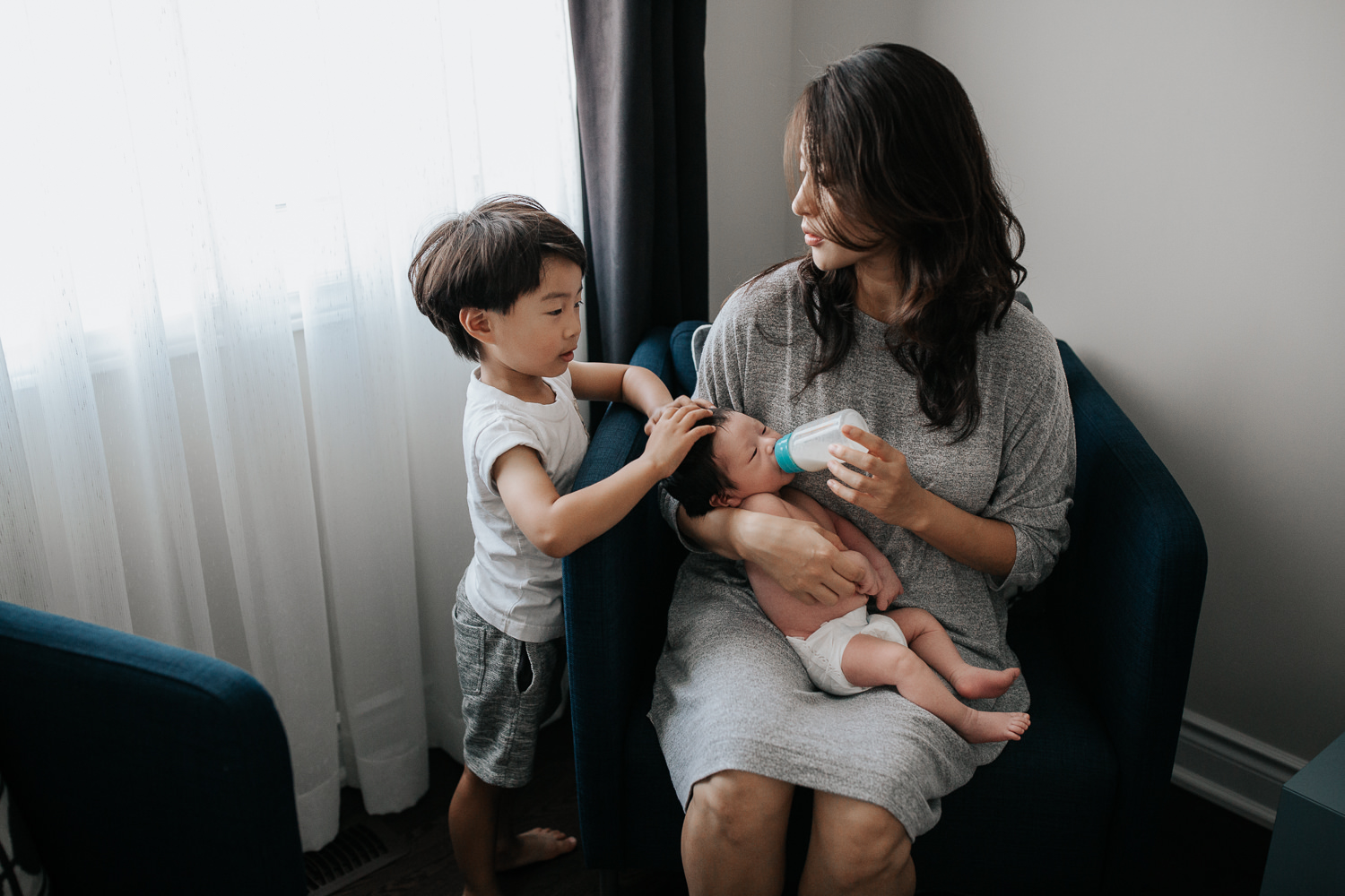 mom with long dark hair sits in blue armchair feeding 2 week old baby boy in diaper a bottle, 3 year old toddler son standing next to her with hands on little brother's head - Markham In-Home Photos