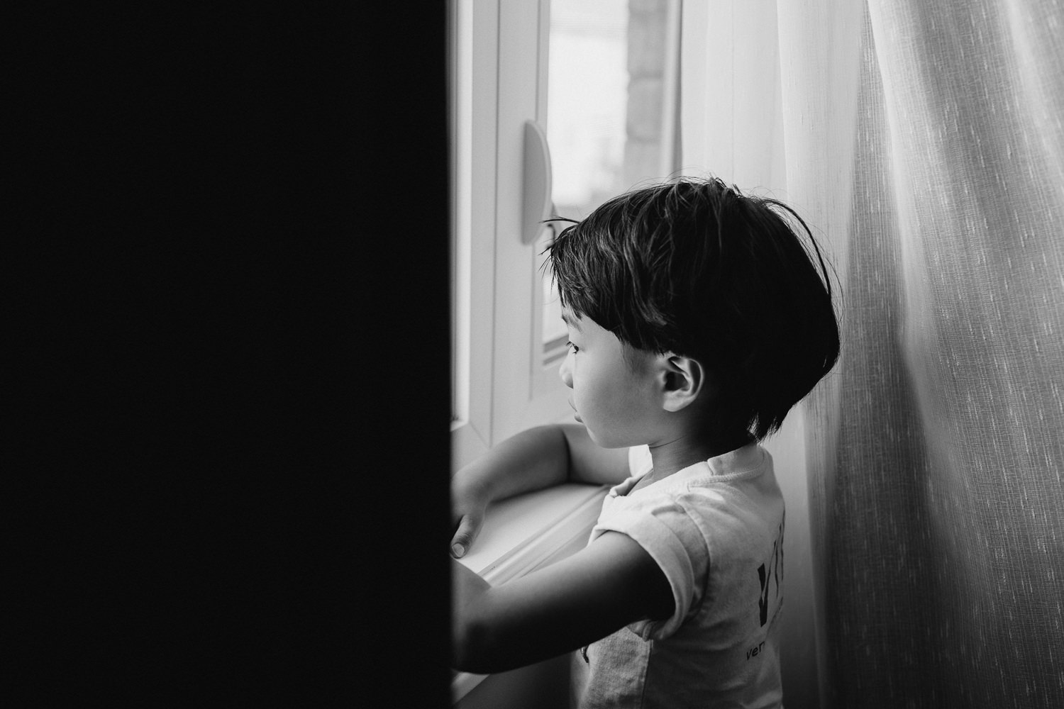 3 year old boy with dark hair and eyes wearing light t-shirt standing at window looking outside - GTA In-Home Photos