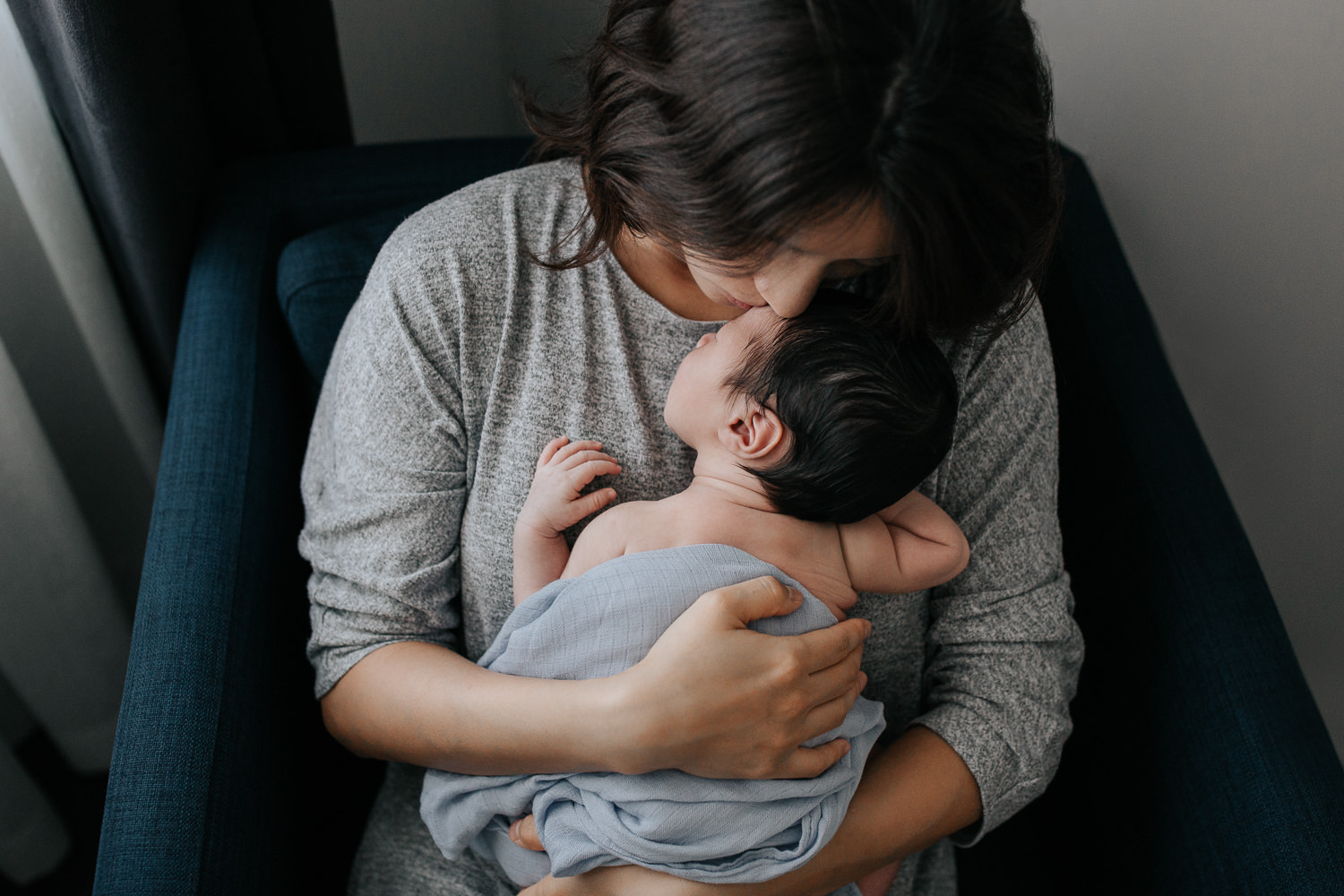 mother with long dark hair sitting in blue armchair snuggling 2 week old sleeping baby boy to her chest, son wrapped in swaddle - York Region In-Home Photos