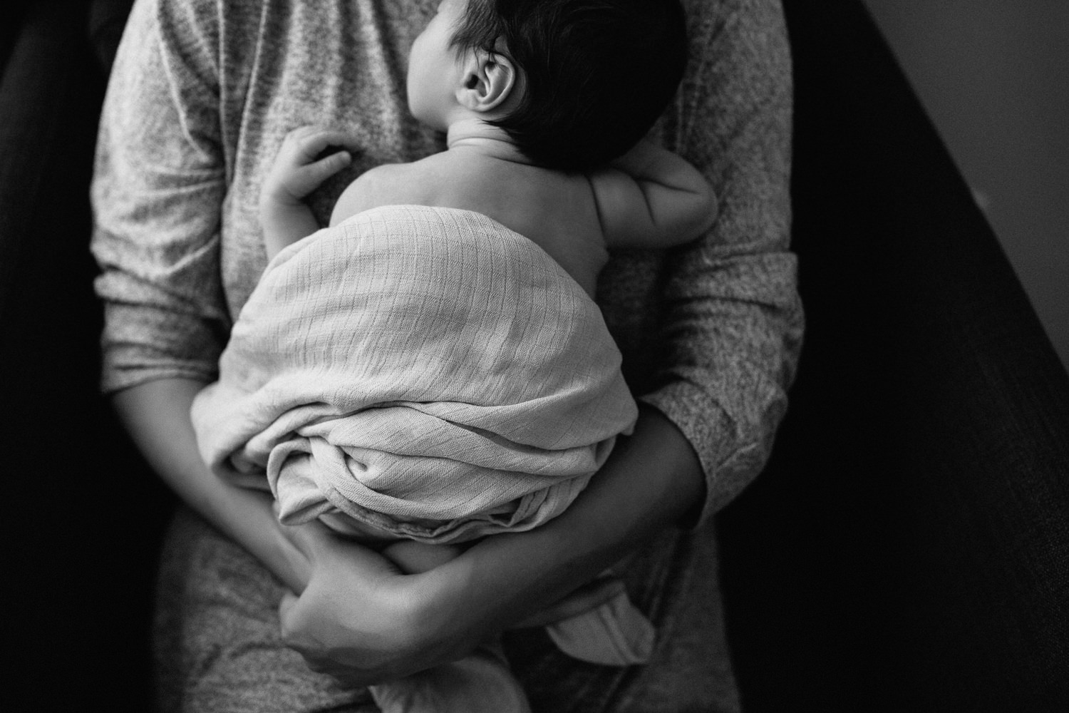 mother sitting in armchair snuggling 2 week old sleeping baby boy to her chest, son wrapped in swaddle - Newmarket Lifestyle Photos