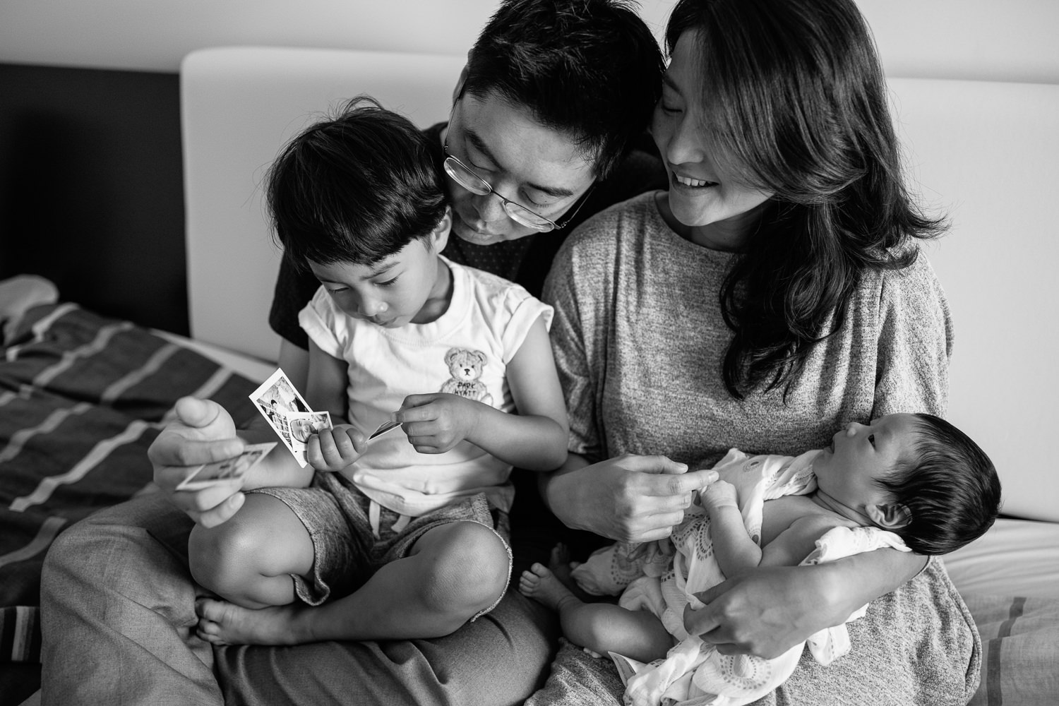 family of 4 sitting on bed, 3 year old toddler boy sitting in dad's lap looking at polaroid pictures in his hands, mom holding 2 week old baby son and sitting next to husband - GTA Lifestyle Photos
