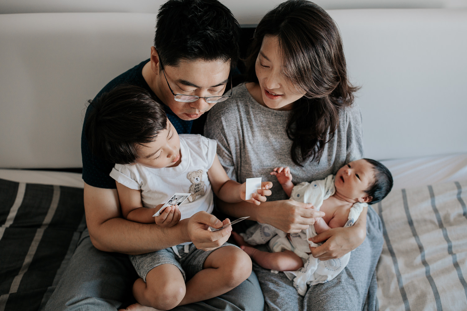 family of 4 sitting on bed, 3 year old toddler boy sitting in dad's lap looking at polaroid pictures in his hands, mom holding 2 week old baby son and sitting next to husband - Barrie Lifestyle Photos