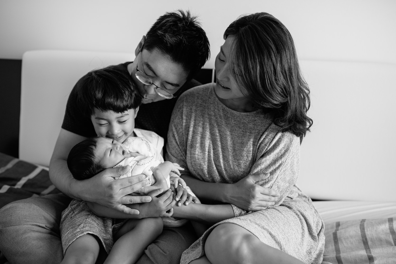 family of 4 sitting on bed, 3 year old toddler boy sitting in dad's lap holding 2 week old baby brother, mom snuggled next to them - Newmarket Lifestyle Photography