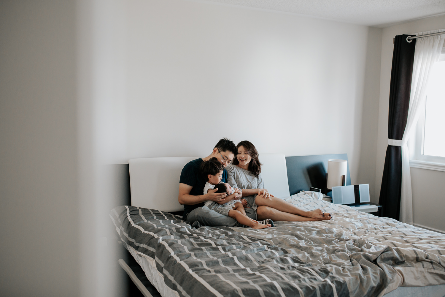 family of 4 sitting on bed, 3 year old toddler boy sitting in dad's lap holding 2 week old baby brother, mom snuggled next to them on the bed - York Region Lifestyle Photos