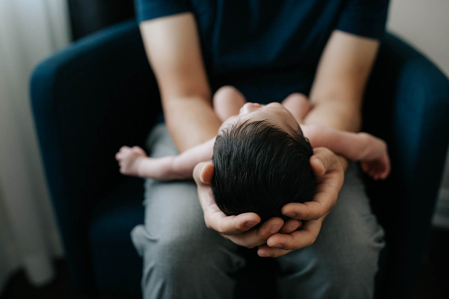 father sitting in navy blue armchair holding sleeping 2 week old baby boy in diaper, son's head in dad's hands, close up of hair - Barrie Lifestyle Photography