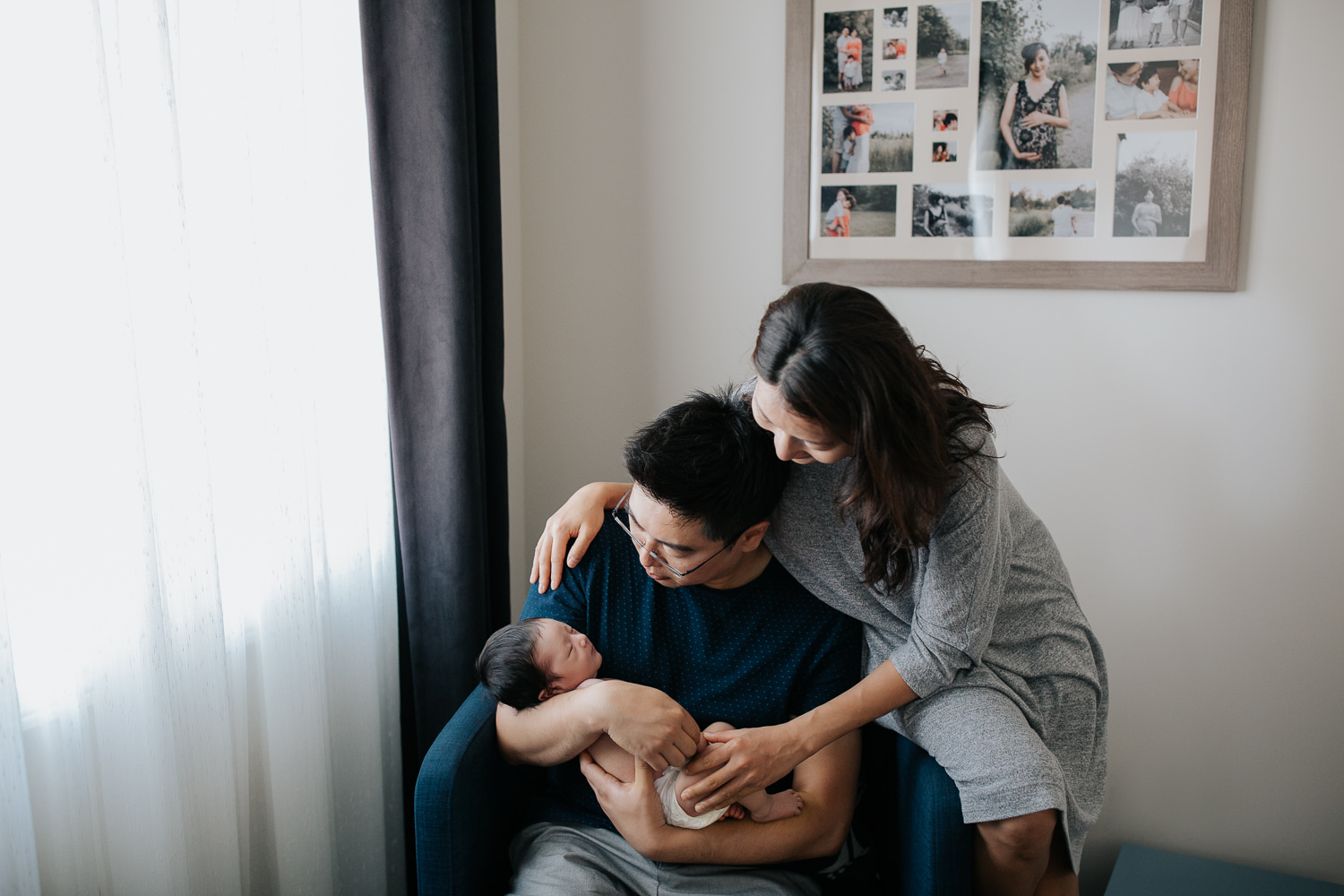 father sitting in navy blue armchair holding sleeping 2 week old baby boy in diaper, mom sitting on chair's edge, arm around husband leaning over to look at son - York Region Lifestyle Photography