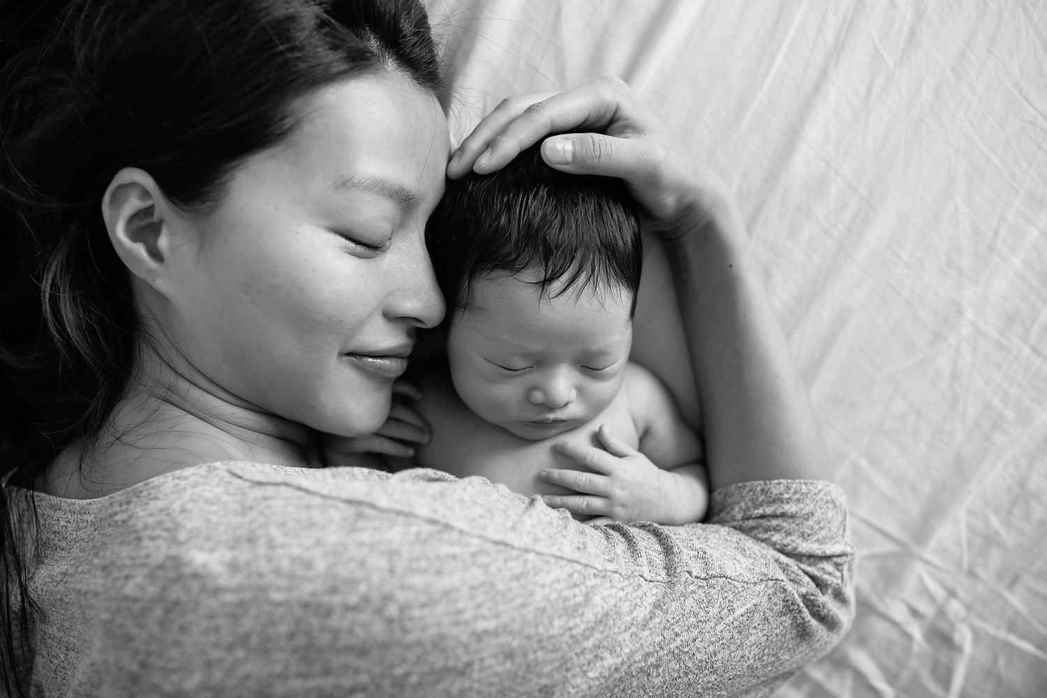 mother with long dark hair lying on bed, sleeping 2 week old baby boy snuggled next to her face, her arm around him and hand resting in son's head, eyes closed - Stouffville Lifestyle Photos