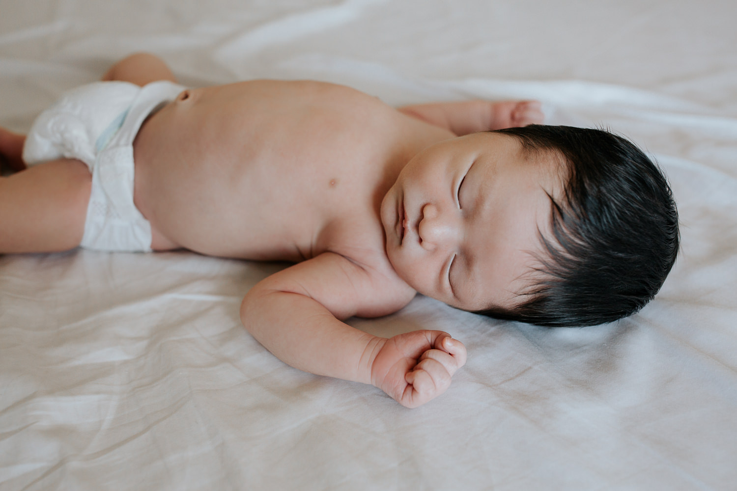 2 week old baby boy with long dark hair in diaper lying on bed, body covered by white swaddle, arms out, head to side, sleeping on bed - Markham Lifestyle Photos