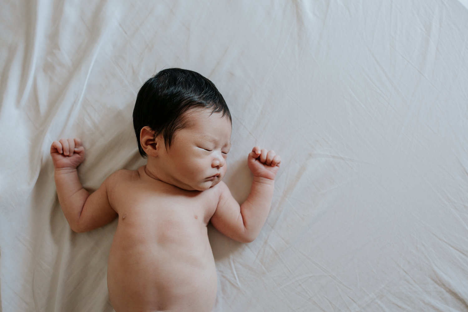 2 week old baby boy with long dark hair in diaper lying on bed, body covered by white swaddle, arms out, head to side, sleeping - Uxbridge Lifestyle Photos
