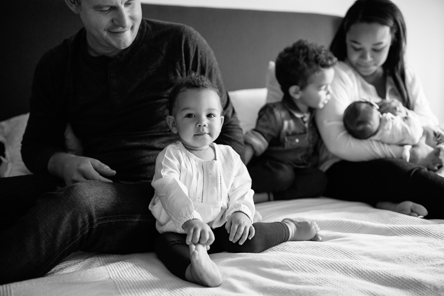 1 year old girl with dark hair and eyes sitting next to dad on master bed looking at camera, mom, brother and baby sister in background - Newmarket Lifestyle Photography