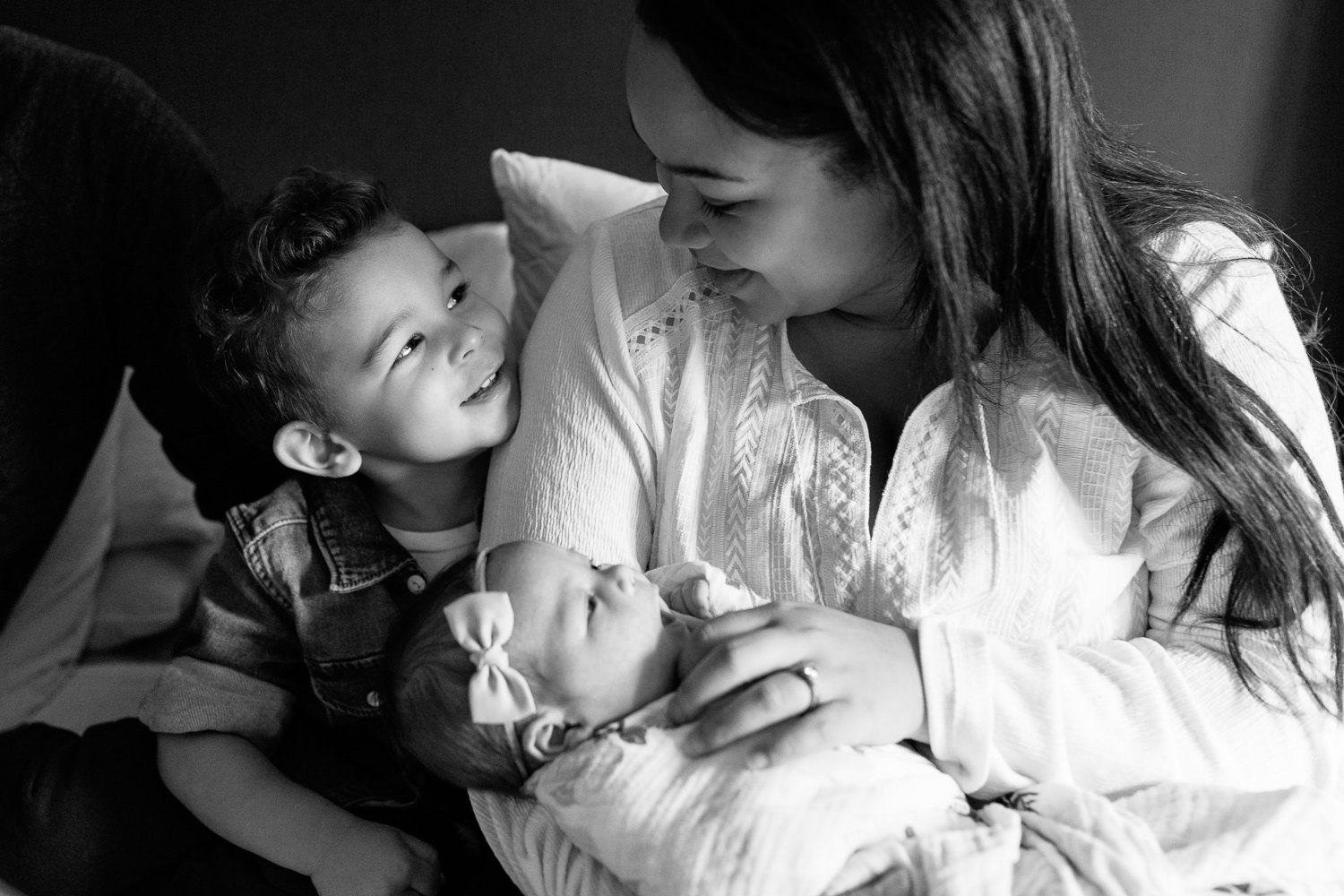 2 year old big brother sitting on master bed next to mom holding 2 week old baby girl, boy smiling up at mom - Markham Lifestyle Photography