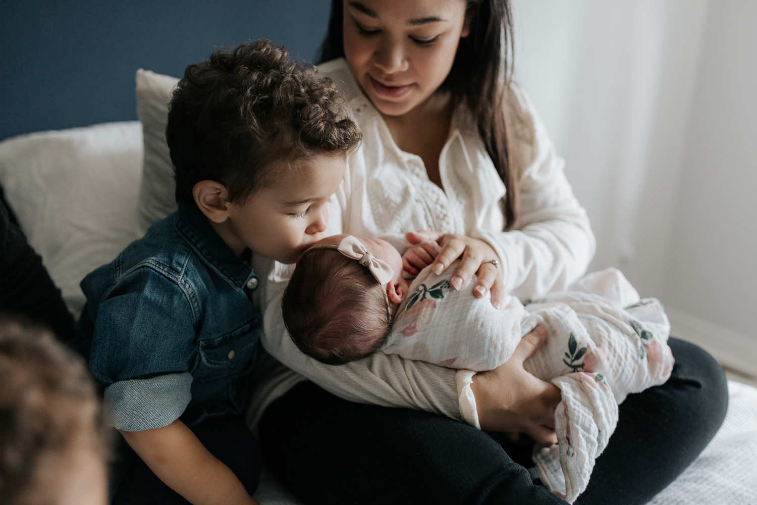 2 year old big brother sitting on master bed next to mom holding 2 week old baby girl, boy kissing little sister on head - Stouffville Lifestyle Photography