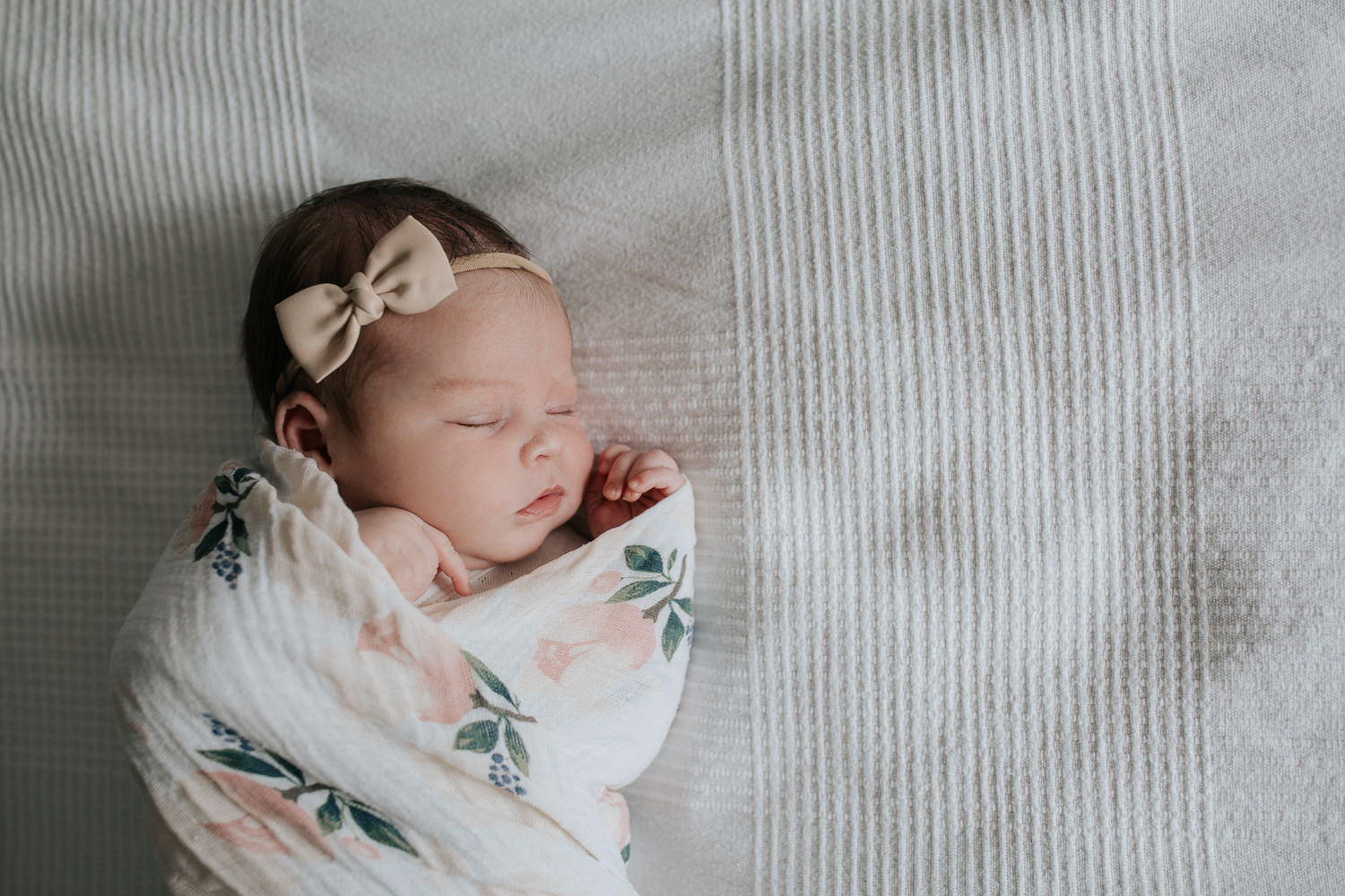 2 week old baby girl with dark hair wrapped in swaddle with soft pink flowers, wearing bow headband asleep on bed, hands near face - Barrie Lifestyle Photography