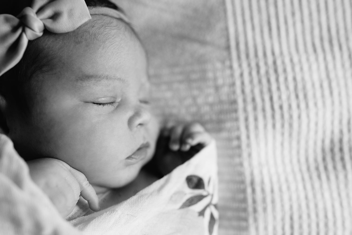 2 week old baby girl with dark hair wrapped in swaddle with flowers, wearing bow headband asleep on bed, hands near face, close up of face - GTA Lifestyle Photography