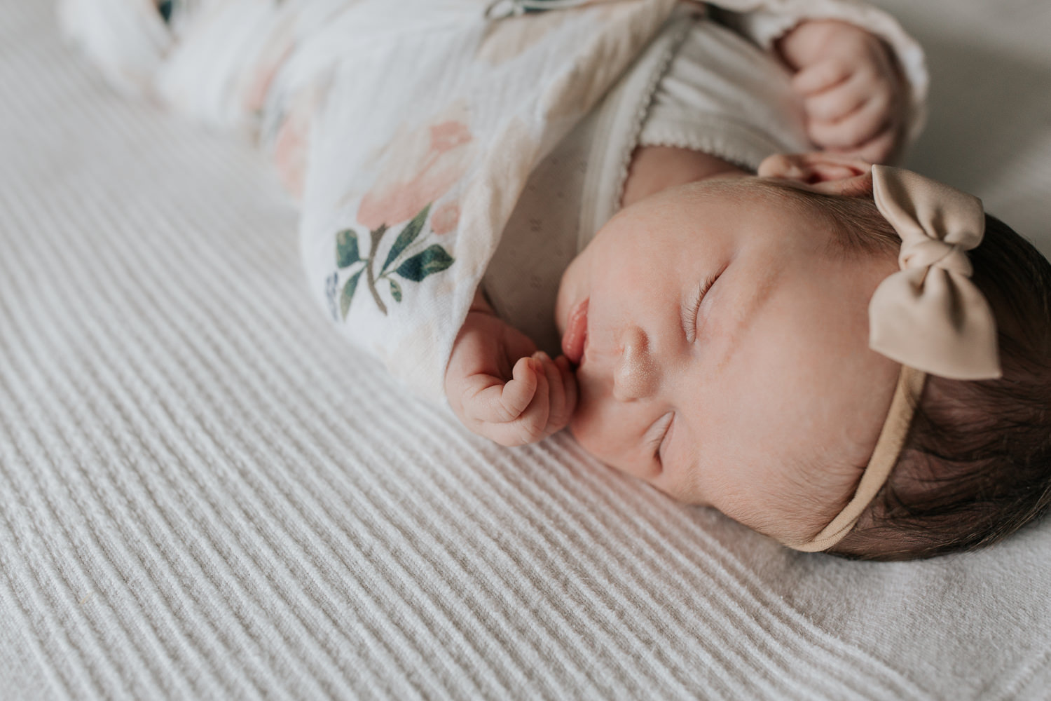2 week old baby girl with dark hair wrapped in swaddle with soft pink flowers, wearing bow headband asleep on bed, hands near face - York Region Lifestyle Photography