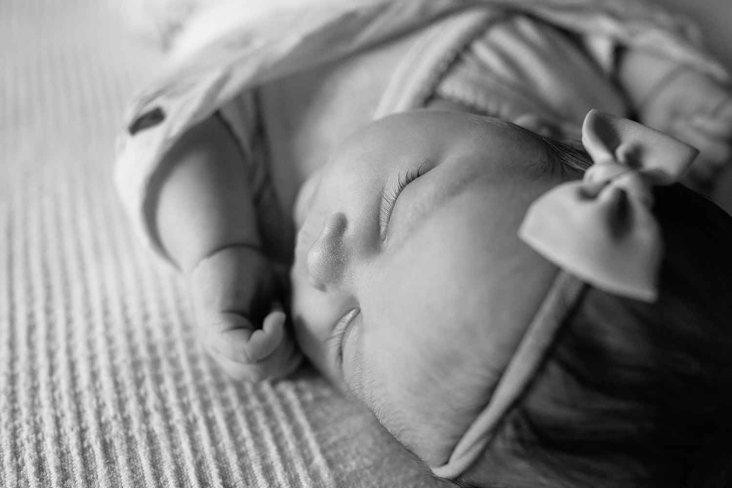 2 week old baby girl with dark hair wrapped in swaddle with flowers, wearing bow headband asleep on bed, hands near face, close up of eyelashes - Newmarket In-Home Photography