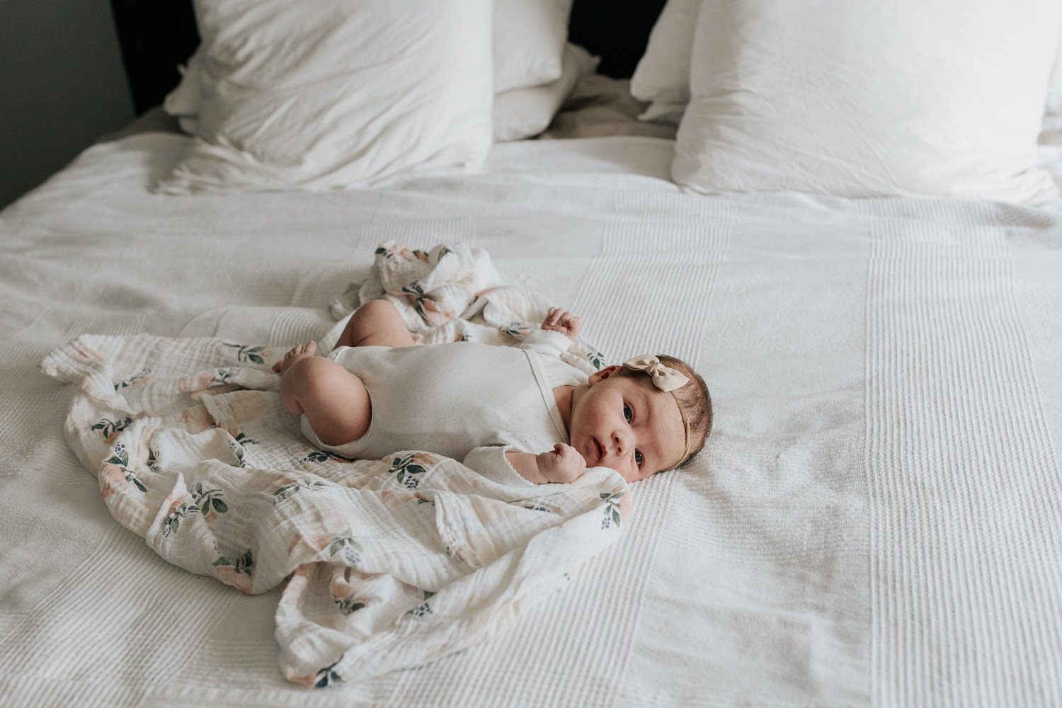 2 week old baby girl with dark hair in white onesie lying on swaddle with soft pink flowers, wearing bow headband awake on bed, looking at camera -​​​​​​​ GTA In-Home Photography