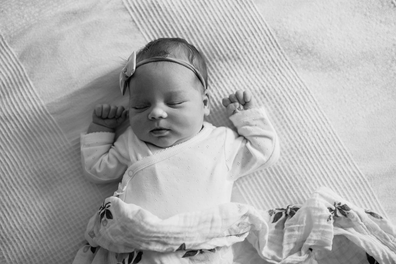 2 week old baby girl with dark hair in white onesie lying on swaddle, wearing bow headband asleep on bed, hands near face -​​​​​​​ York Region In-Home Photography