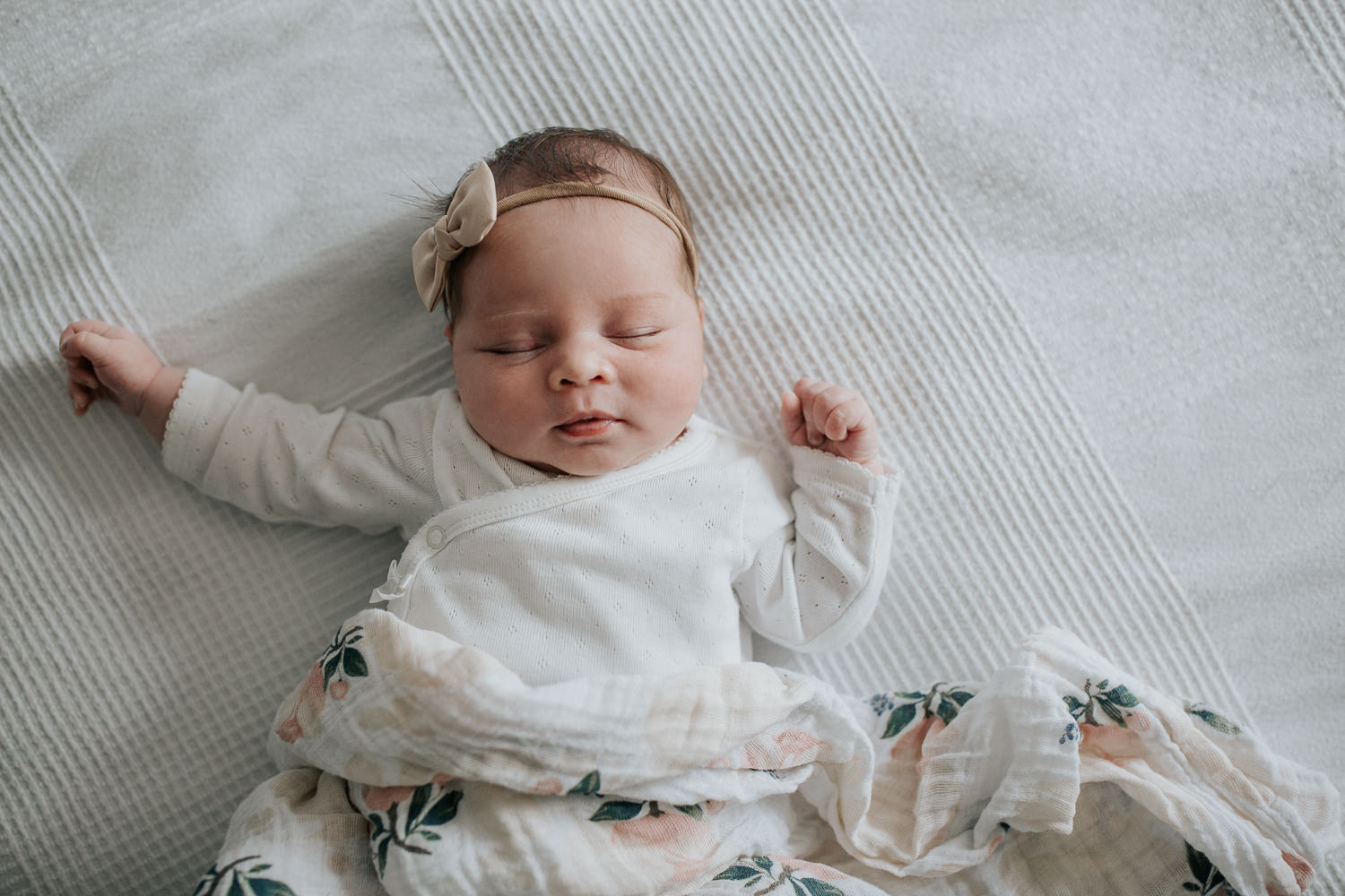 2 week old baby girl with dark hair in white onesie lying on swaddle with soft pink flowers, wearing bow headband asleep on bed, hands near face -​​​​​​​ Newmarket In-Home Photos