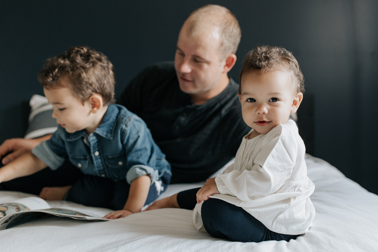 father sitting on bed with 2 year old son reading a story, 1 year old daughter sitting next to them, looking over her shoulder at camera - Markham In-Home Photos