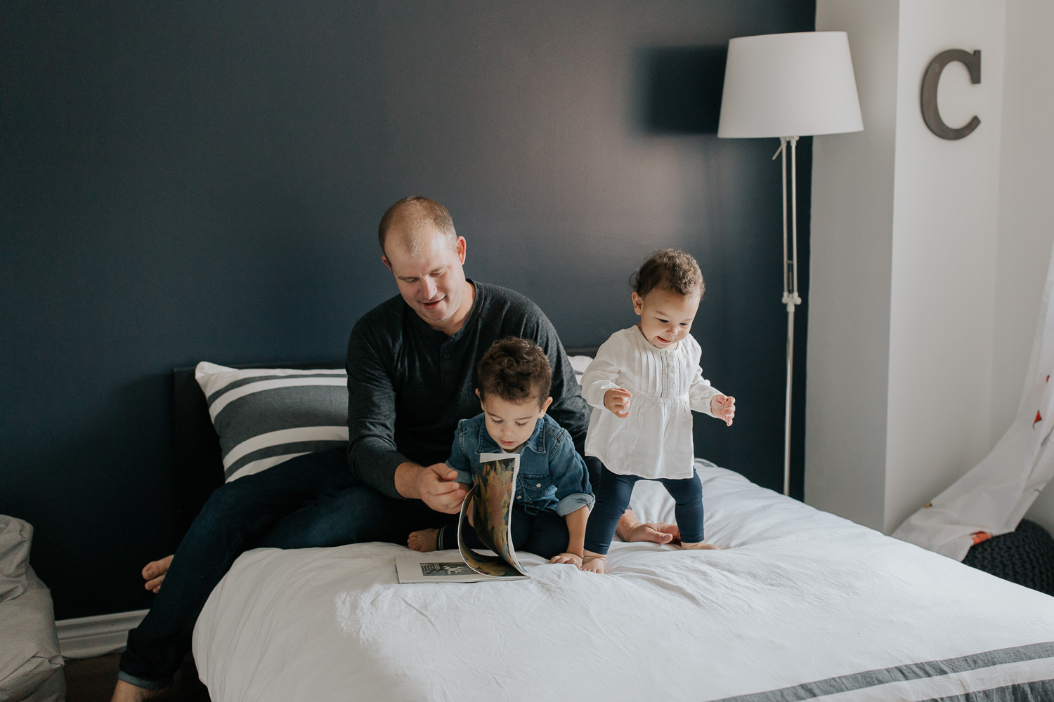father sitting on bed with 2 year old son reading a story, 1 year old daughter standing next to them, jumping - Stouffville In-Home Photos