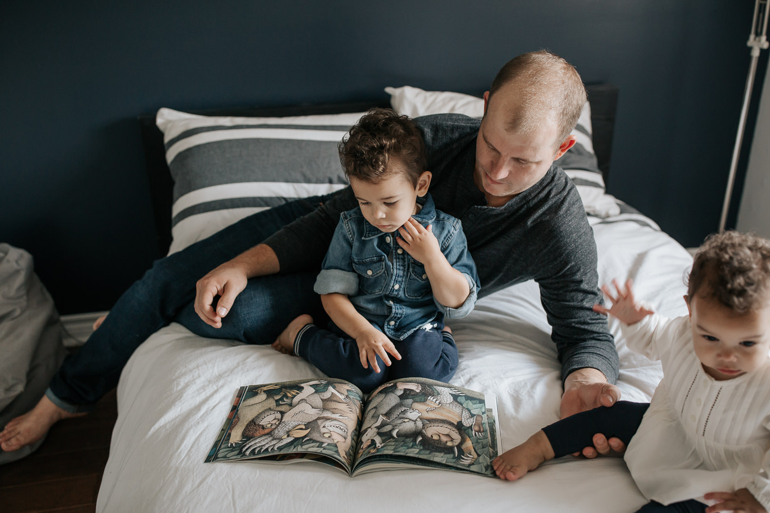 father sitting on bed with 2 year old son reading a story, 1 year old daughter sitting next to them, dad's hand holding her leg - GTA In-Home Photos