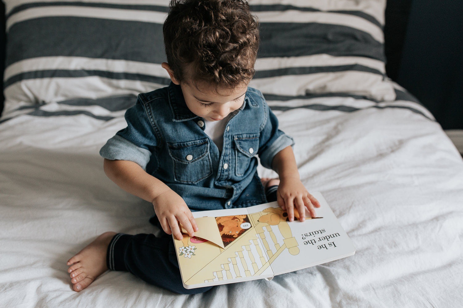 2 year old toddler boy with dark curly hair wearing chambray shirt sitting on his bed reading book - York Region In-Home Photos