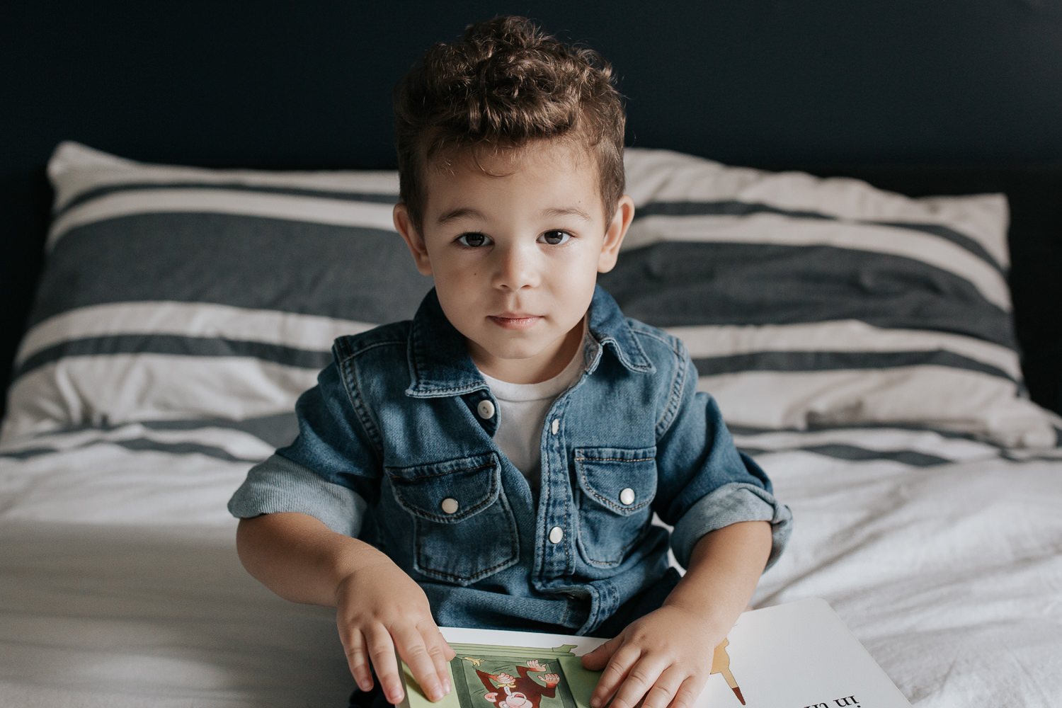 2 year old toddler boy with dark curly hair wearing chambray shirt sitting on his bed holding book and looking at camera - Newmarket Lifestyle Photos