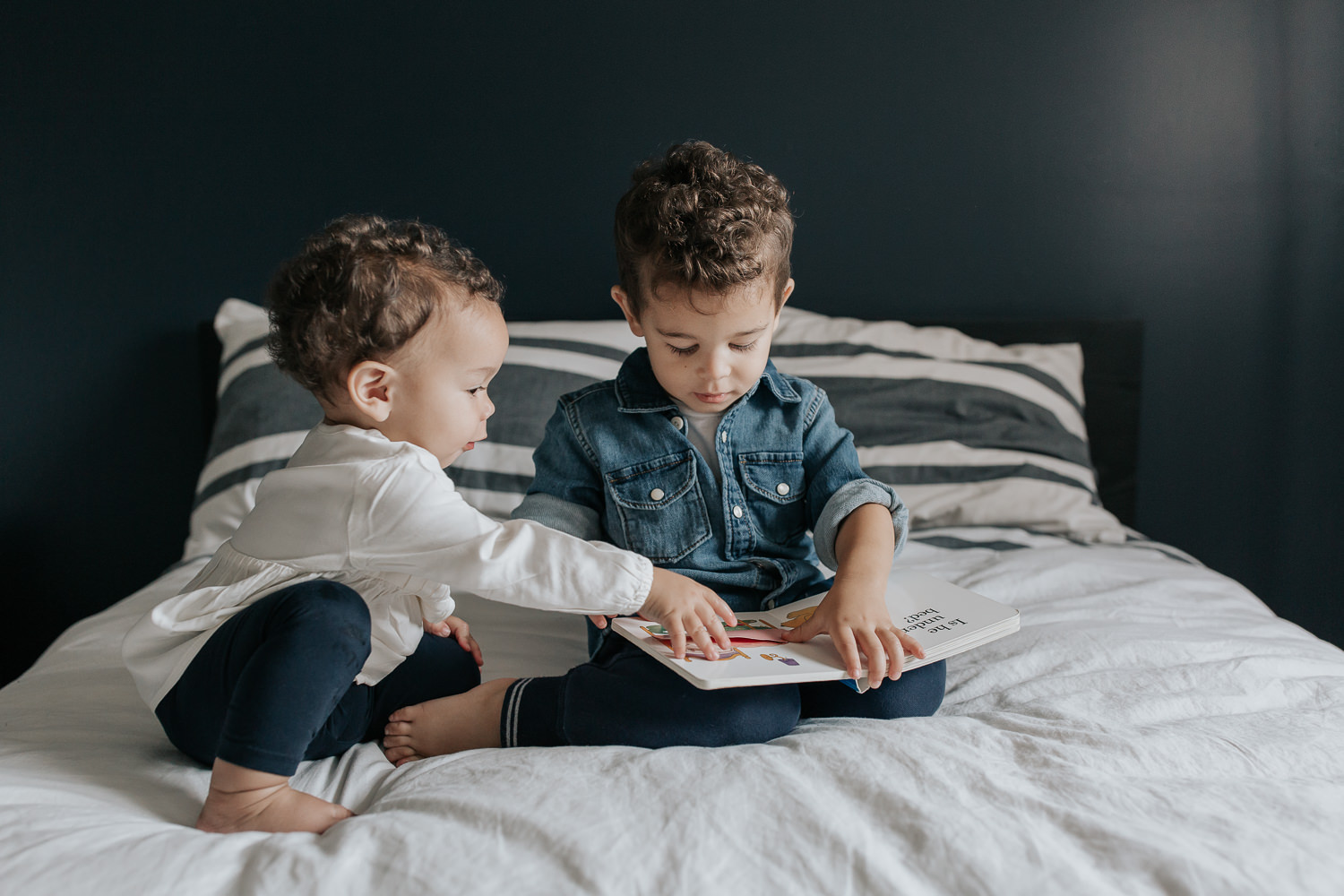 2 year old toddler boy with dark curly hair wearing chambray shirt sitting on his bed reading book, 1 year old baby sister sitting next to him, reaching for the page - Markham Lifestyle Photos