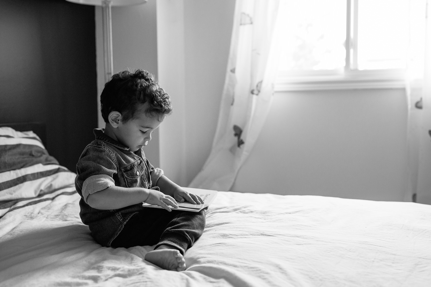 2 year old toddler boy with dark curly hair wearing chambray shirt sitting on his bed reading book - Stouffville Lifestyle Photos