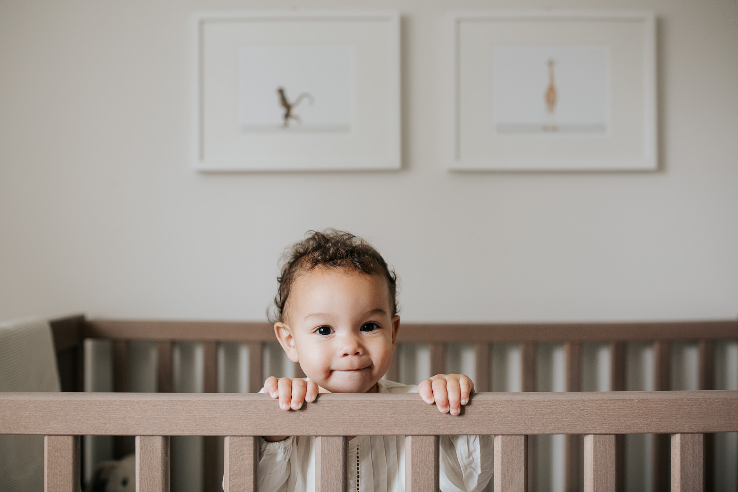 2 year old girl with dark curly hair and brown eyes in cream top and dark pants standing in crib looking at camera in neutral nursery with animal prints on wall - Newmarket Lifestyle Photography