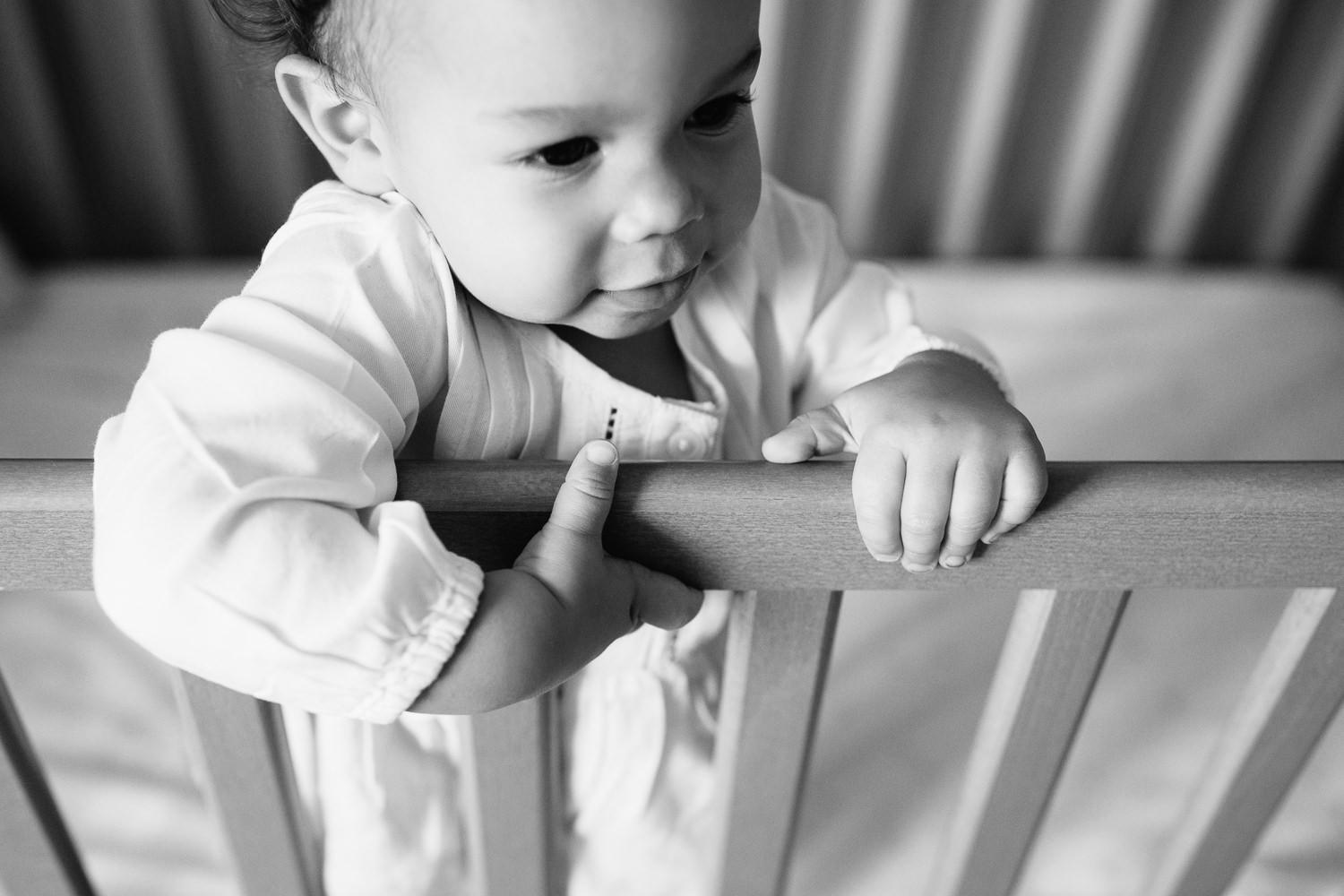 2 year old girl with dark curly hair and brown eyes in light top standing in nursery crib, close up of hands holding onto rail - Stouffville Lifestyle Photography