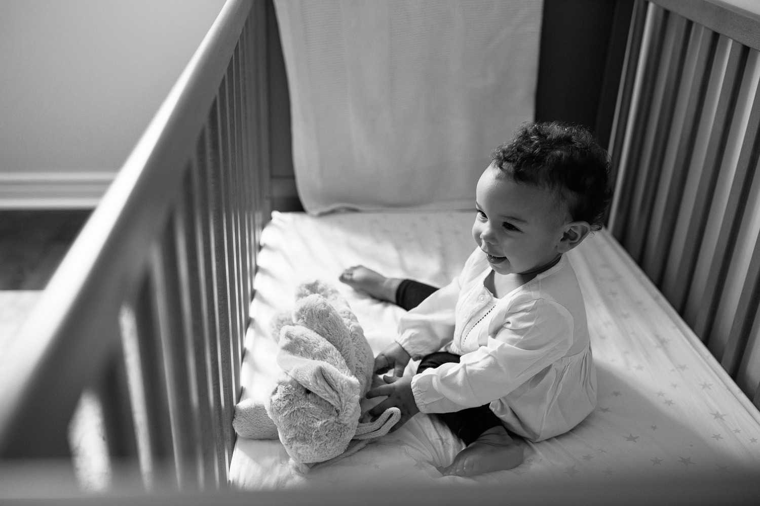 2 year old girl with dark curly hair and brown eyes in cream top and dark pants sitting in crib holding stuffed bunny and smiling - York Region Lifestyle Photos