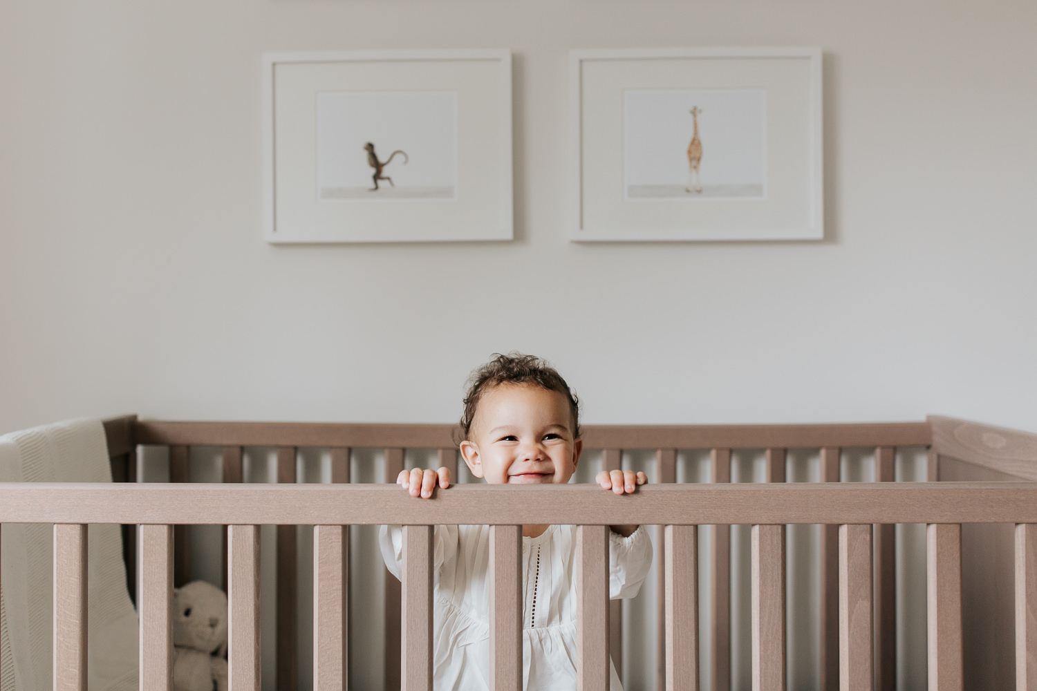 2 year old girl with dark curly hair and brown eyes in cream top and dark pants standing in crib looking up and smiling in neutral nursery with animal prints on wall - Markham Lifestyle Photography