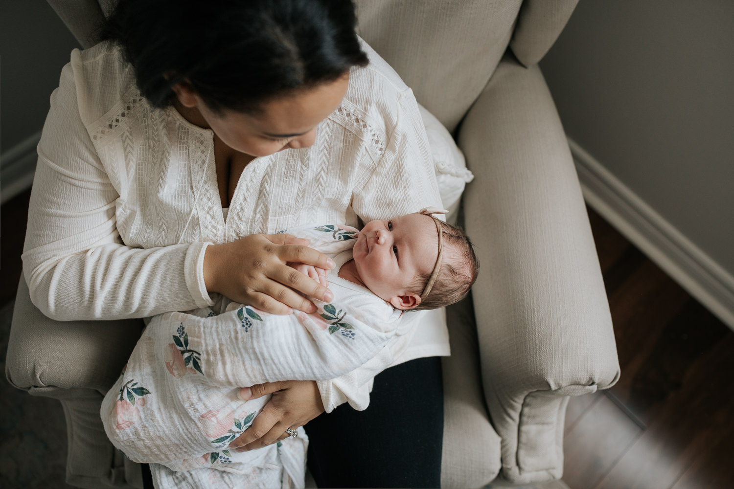 mother sitting in nursery glider chair holding and looking at 2 week old daughter wrapped in pink floral swaddle with headband on, baby girl looking up at mom - York Region Lifestyle Photography