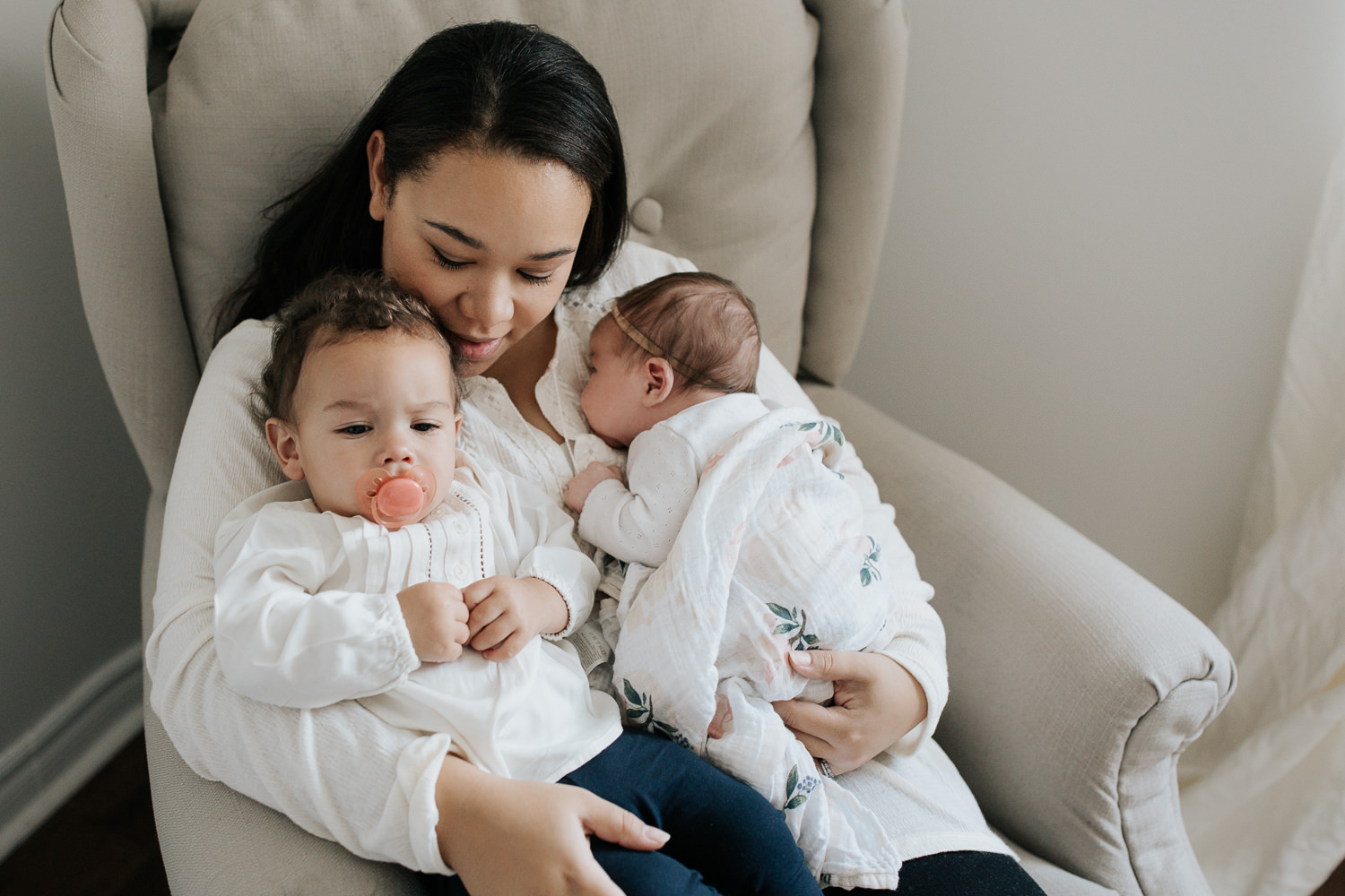 mother sitting in nursery glider chair holding 2 week old baby girl wrapped in swaddle lying against her chest and 1 year old daughter sitting in lap, cuddling - Newmarket In-Home Photography