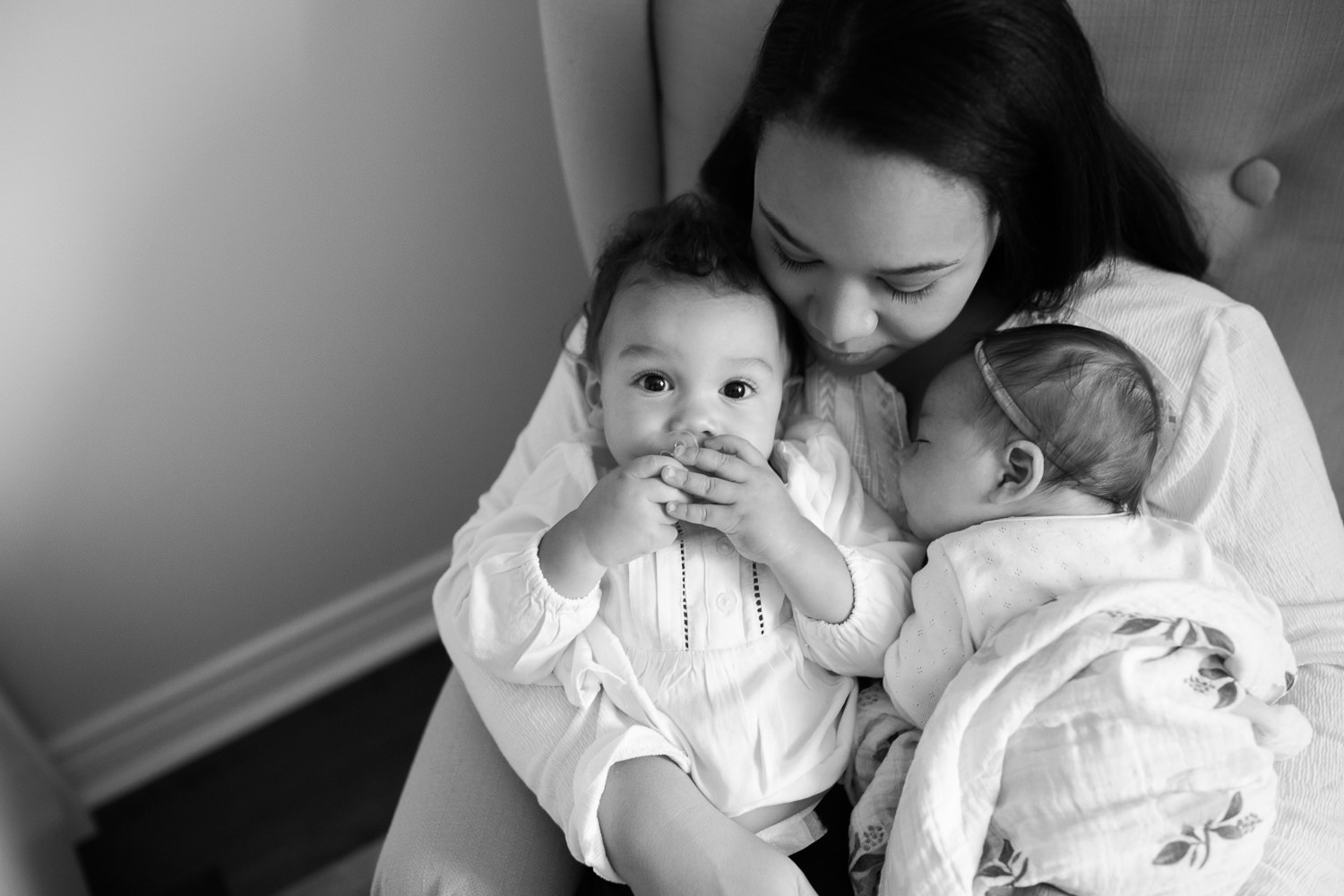 mother sitting in nursery glider chair holding 2 week old baby girl wrapped in swaddle lying against her chest and 1 year old daughter sitting in lap, cuddling - Stouffville In-Home Photography