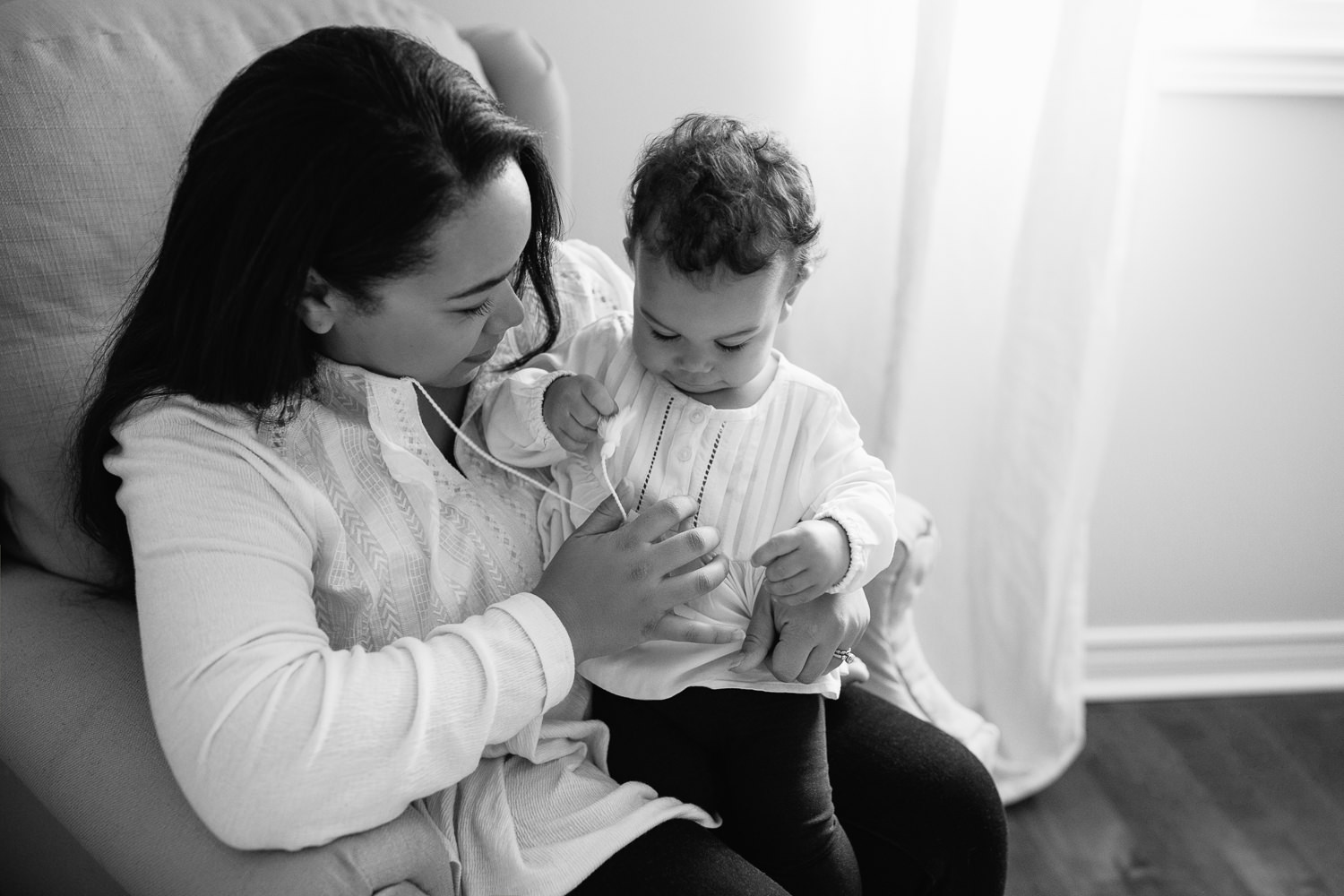 mom with long dark hair in peasant top sitting on nursery glider chair with 1 year old toddler girl with dark curly hair sitting in her lap, playing with strings - York Region In-Home Photography