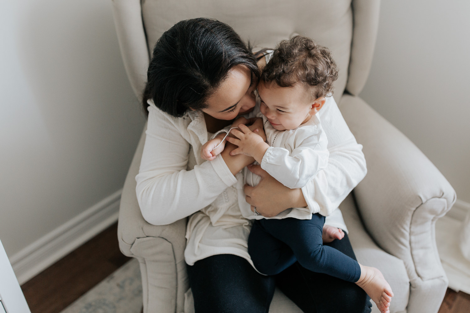 mom with long dark hair in peasant top sitting on nursery glider chair with 1 year old toddler girl with dark curly hair sitting in her lap, mother snuggling daughter - GTA In-Home Photography
