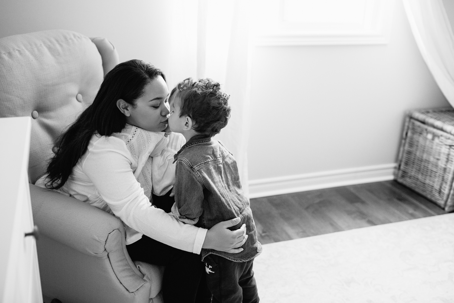 mom sitting on nursery glider chair with 1 year old toddler girl sitting in her lap, 2 year old son standing next to chair and leaning in to give mother a kiss - Newmarket In-Home Photos