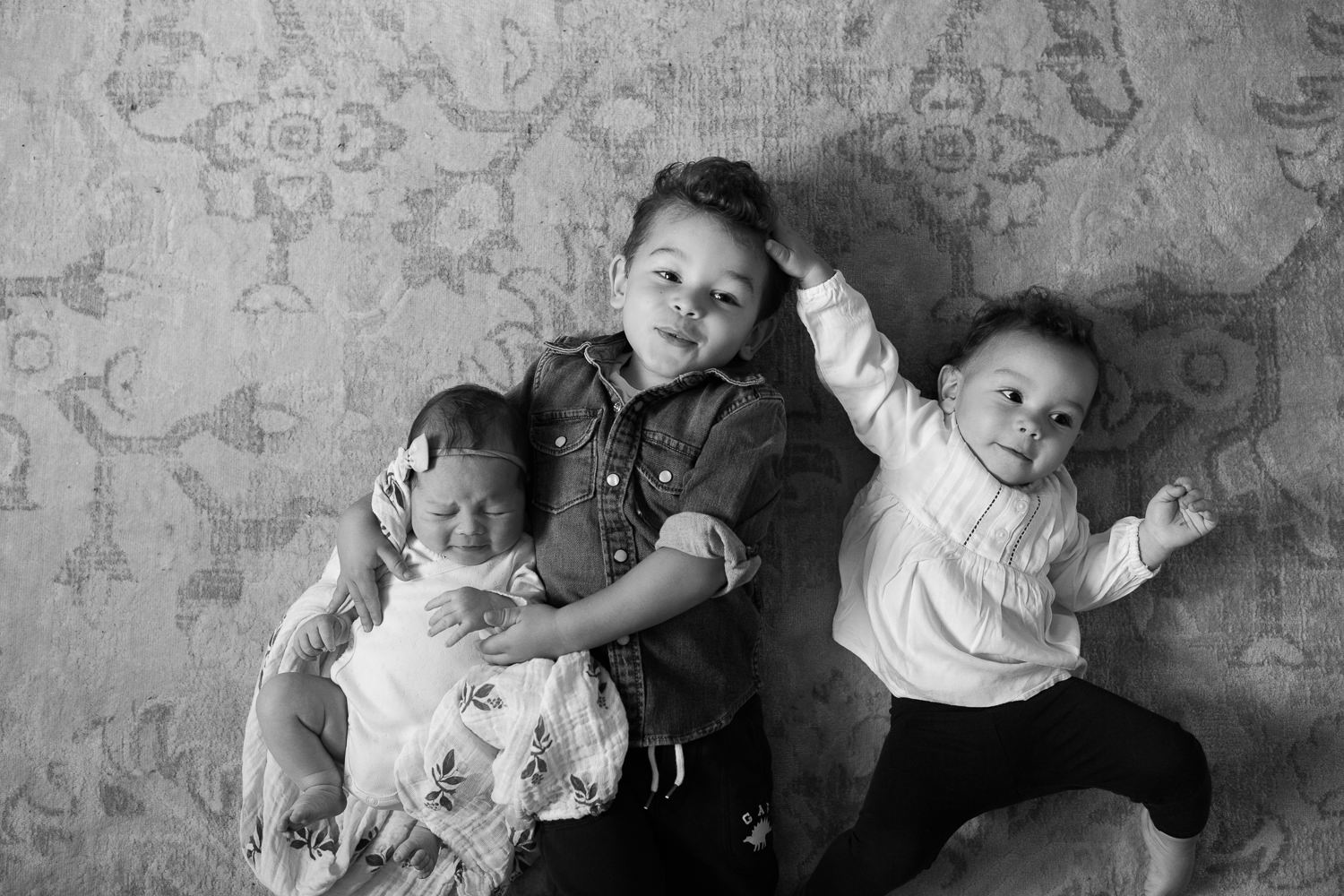 3 siblings lying on floor, 2 week old baby girl held by 2 year old big brother, 1 year old toddler sister next to boy, hand on his head - Markham In-Home Photos