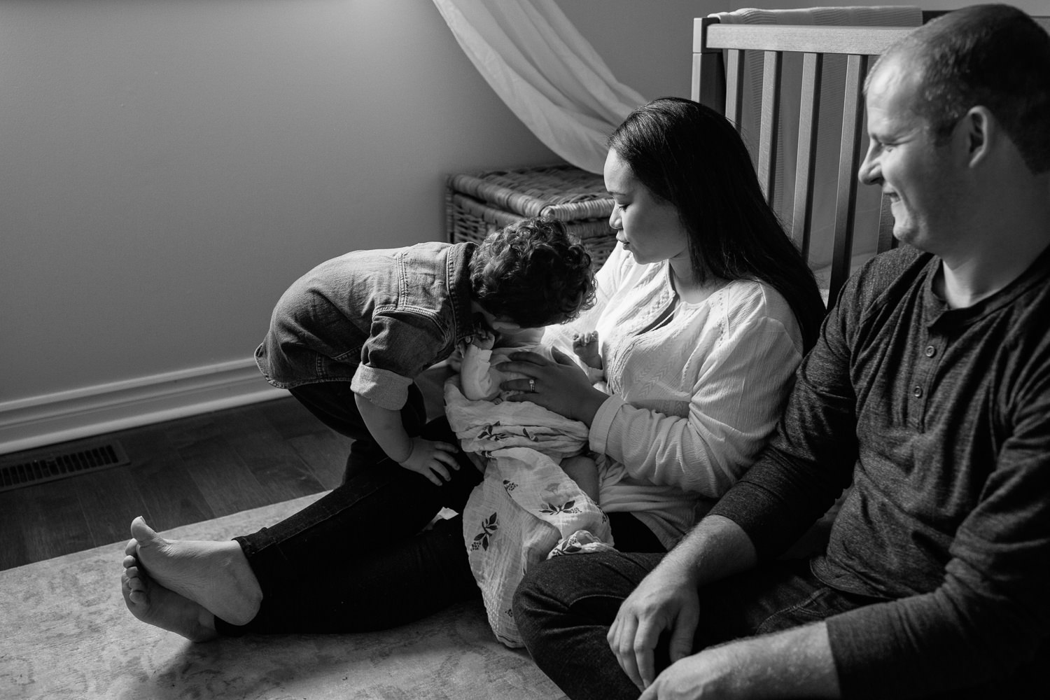 parents sitting on floor of nursery leaning against crib, mom holding 2 week old baby girl in her arms as two year old big brother leans over and kisses little sister - GTA In-Home Photos