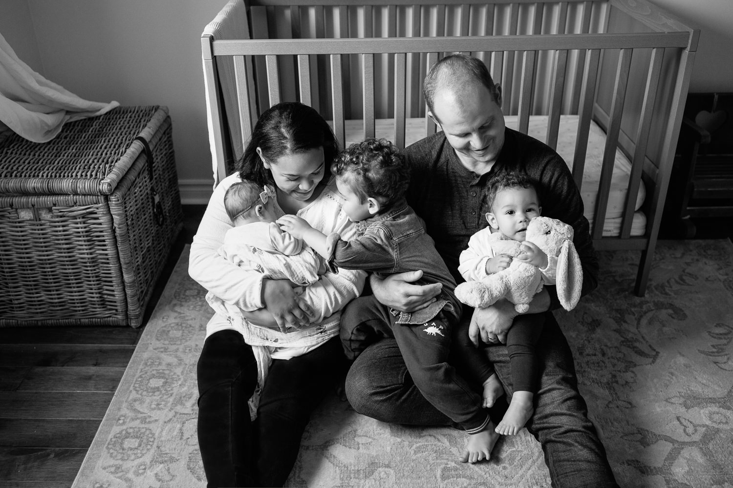 family of 5 sitting on floor of nursery leaning against crib, mom holding 2 week old baby girl, dad snuggling 2 year old boy and 1 year old daughter in his lap - Newmarket Lifestyle Photos