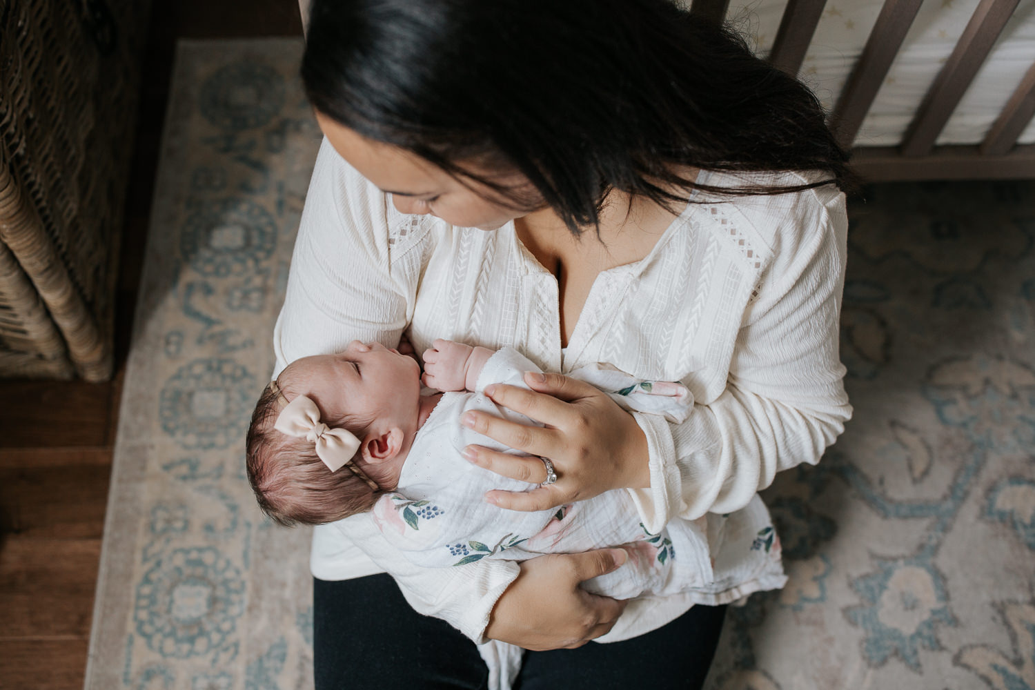 mom with long dark hair sitting on nursery floor leaning against crib holding 2 week old baby girl in swaddle wearing headband as she sleeps in mother's arms - Stouffville Lifestyle Photography