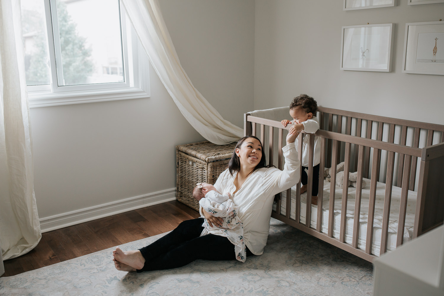 mom with long dark hair sitting on nursery floor holding 2 week old baby girl, smiling and reaching out to 1 year old toddler daughter standing in crib behind her  - Newmarket Lifestyle Photography